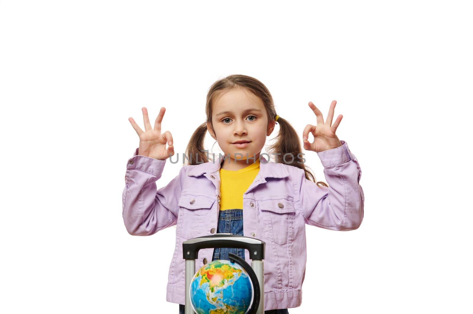 Adorable little traveler girl with two ponytails, standing next to the globe on yellow suitcase baggage, gesturing with hands, demonstrating OK sign looking at camera, isolated on white background