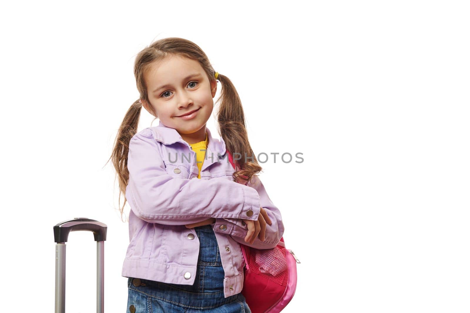 Caucasian preschooler lovely little traveler girl with two ponytails, dressed in purple jacket and blue denim overalls, smiling at camera, posing with pink backpack and suitcase on white background.