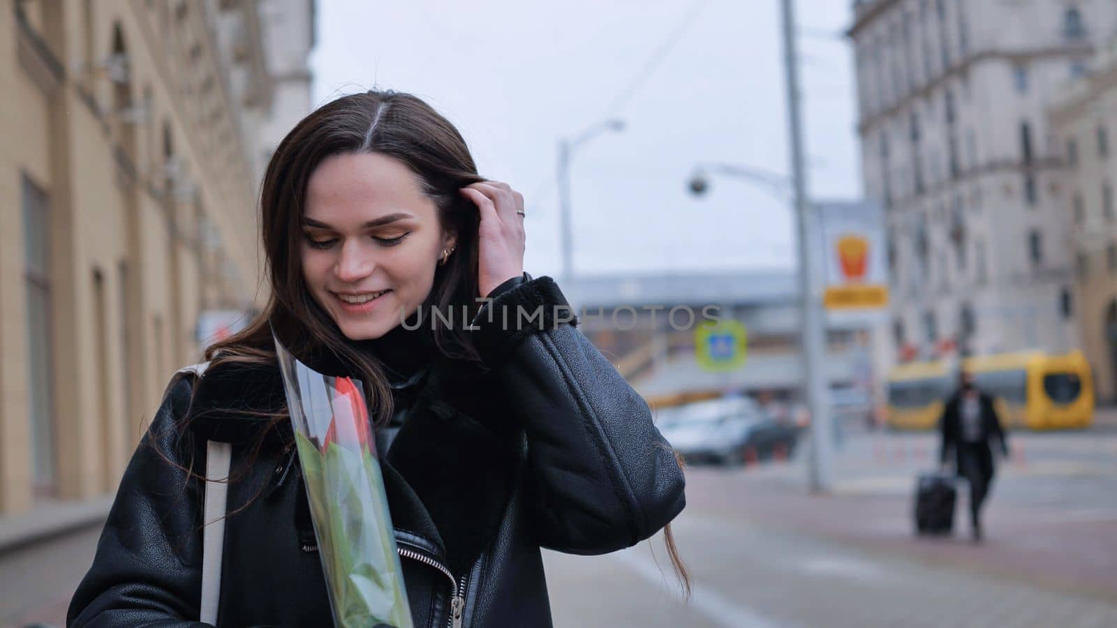 A happy brunette girl in a black jacket walks through the city in early spring