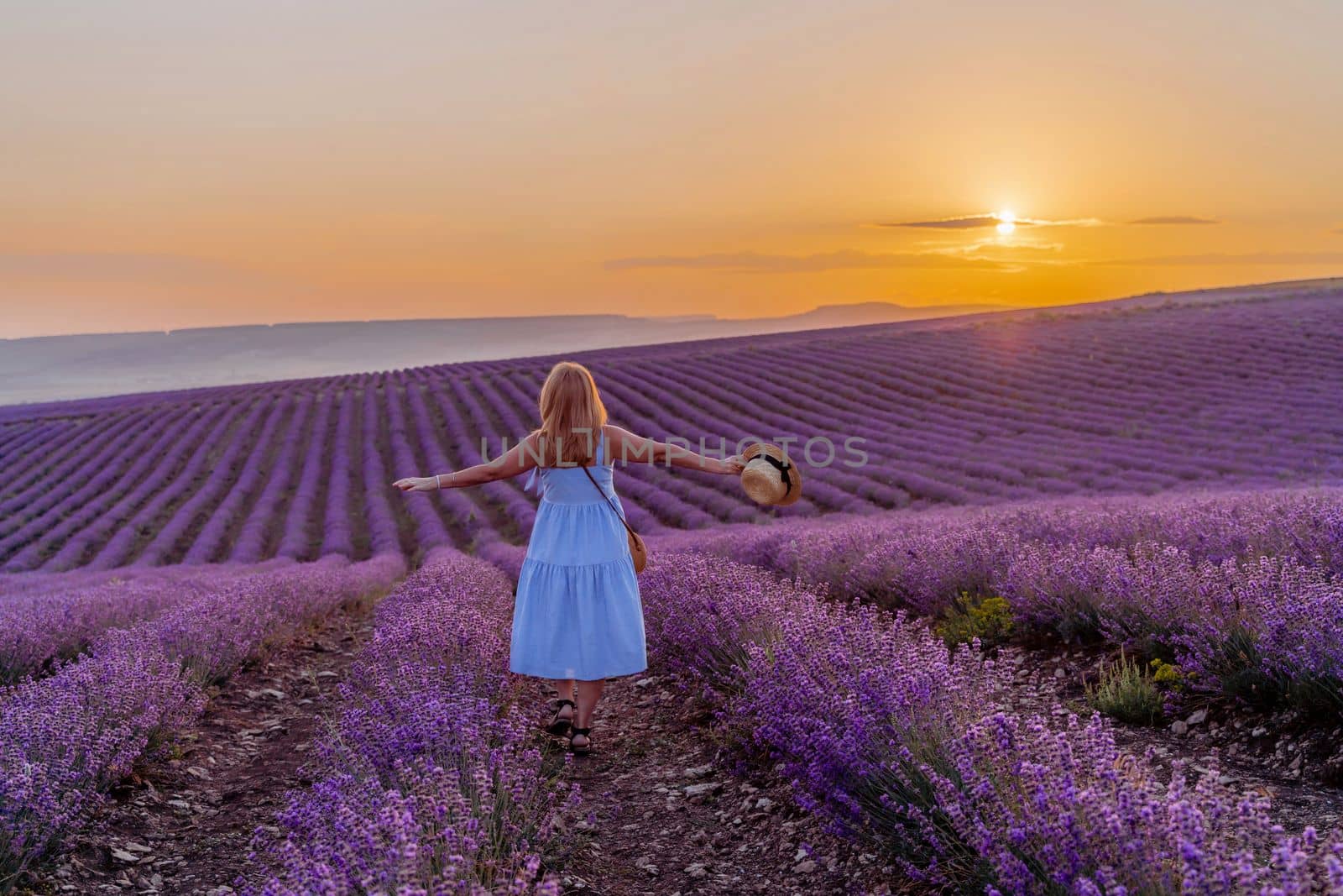 Woman lavender field sunset. Romantic woman walks through the lavender fields. illuminated by sunset sunlight. She is wearing a blue dress with a hat