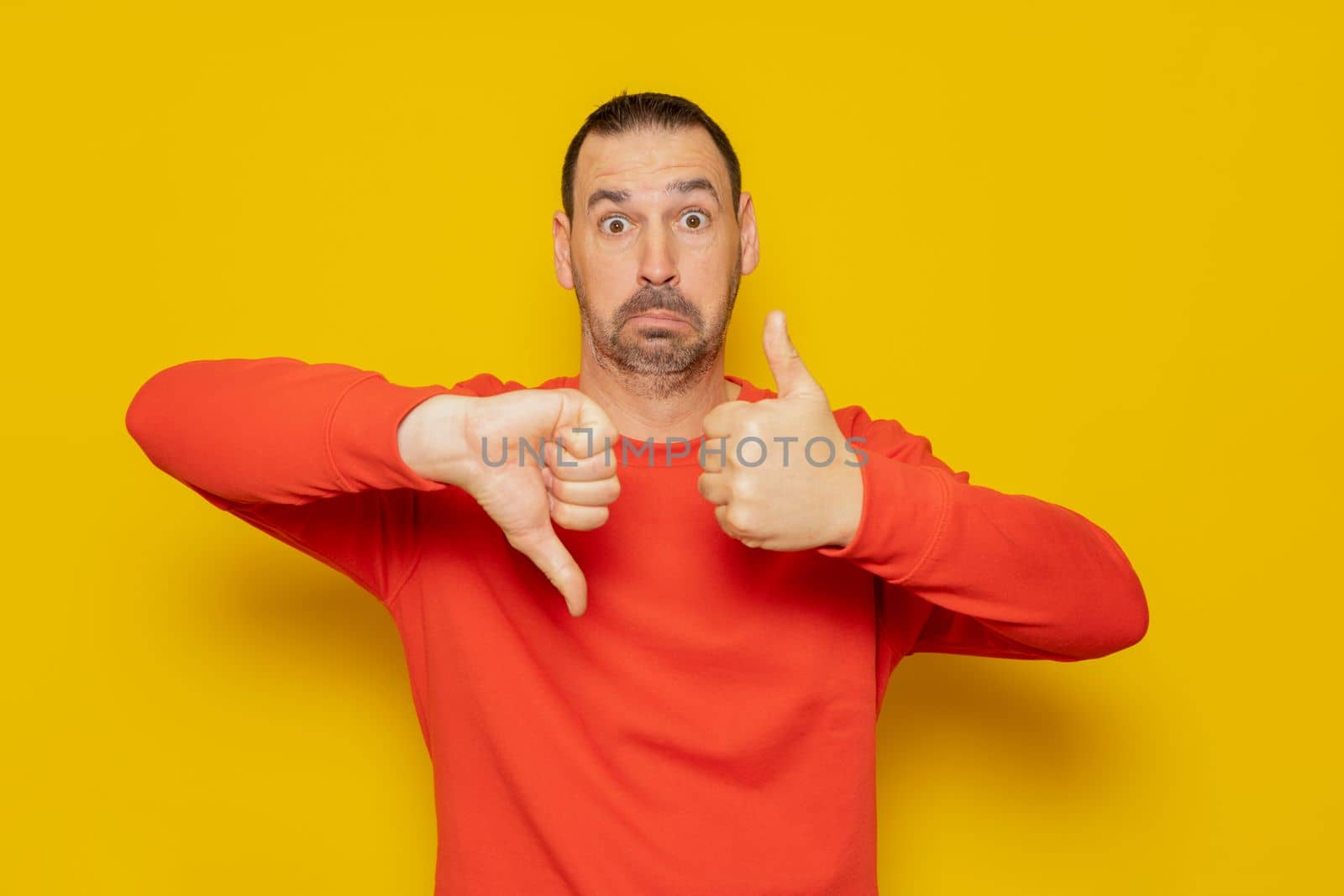 Hispanic man with beard making good and bad signs. Person undecided between yes or no on isolated yellow background