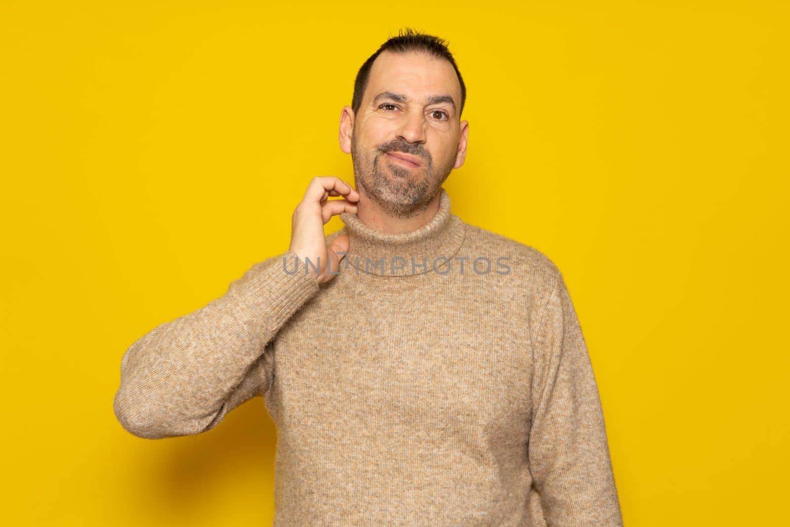 Bearded hispanic man wearing beige turtleneck scratching his neck and grimacing isolated over yellow background. Concept of doubt and indecision