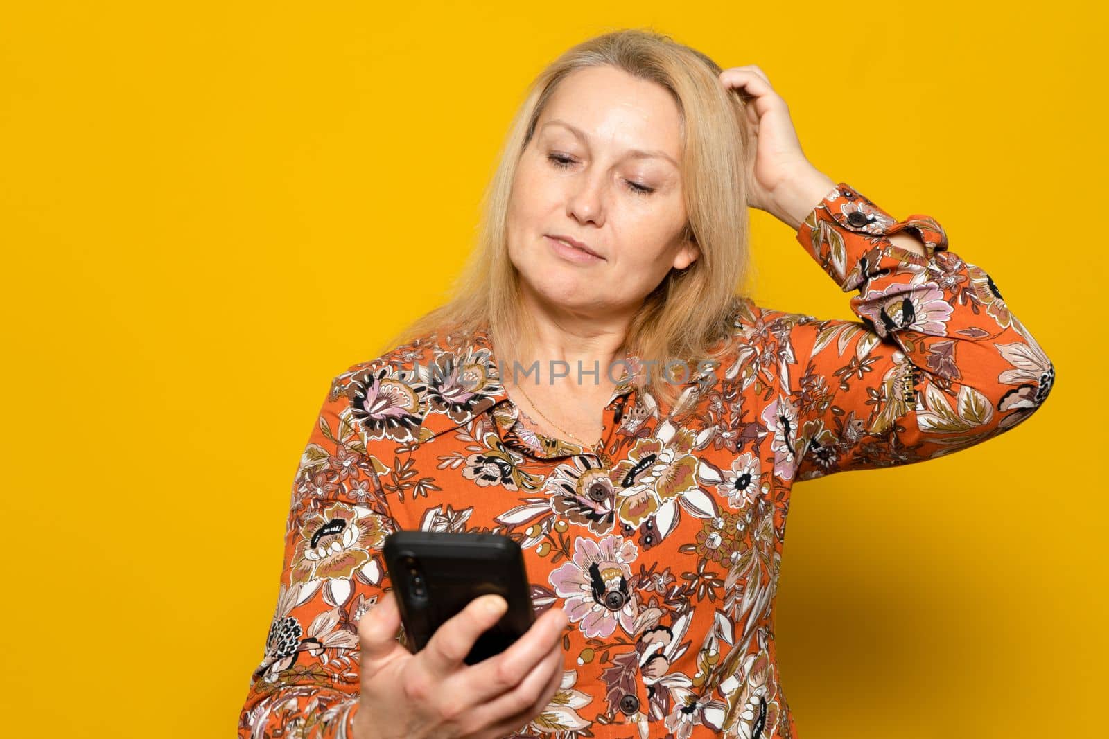 What to text. Portrait of pensive woman holding mobile phone and looking away scratching head with hand isolated over yellow studio background, thinking about question.