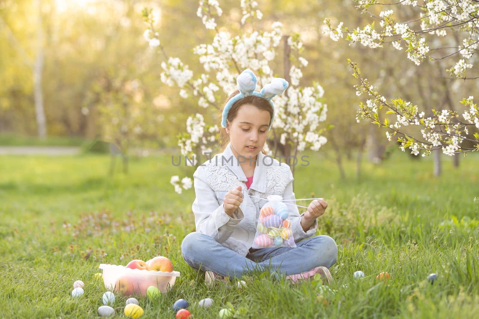 Adorable little girl in bunny ears, blooming tree branch outdoors on a spring day. Kid having fun on Easter egg hunt in the garden.