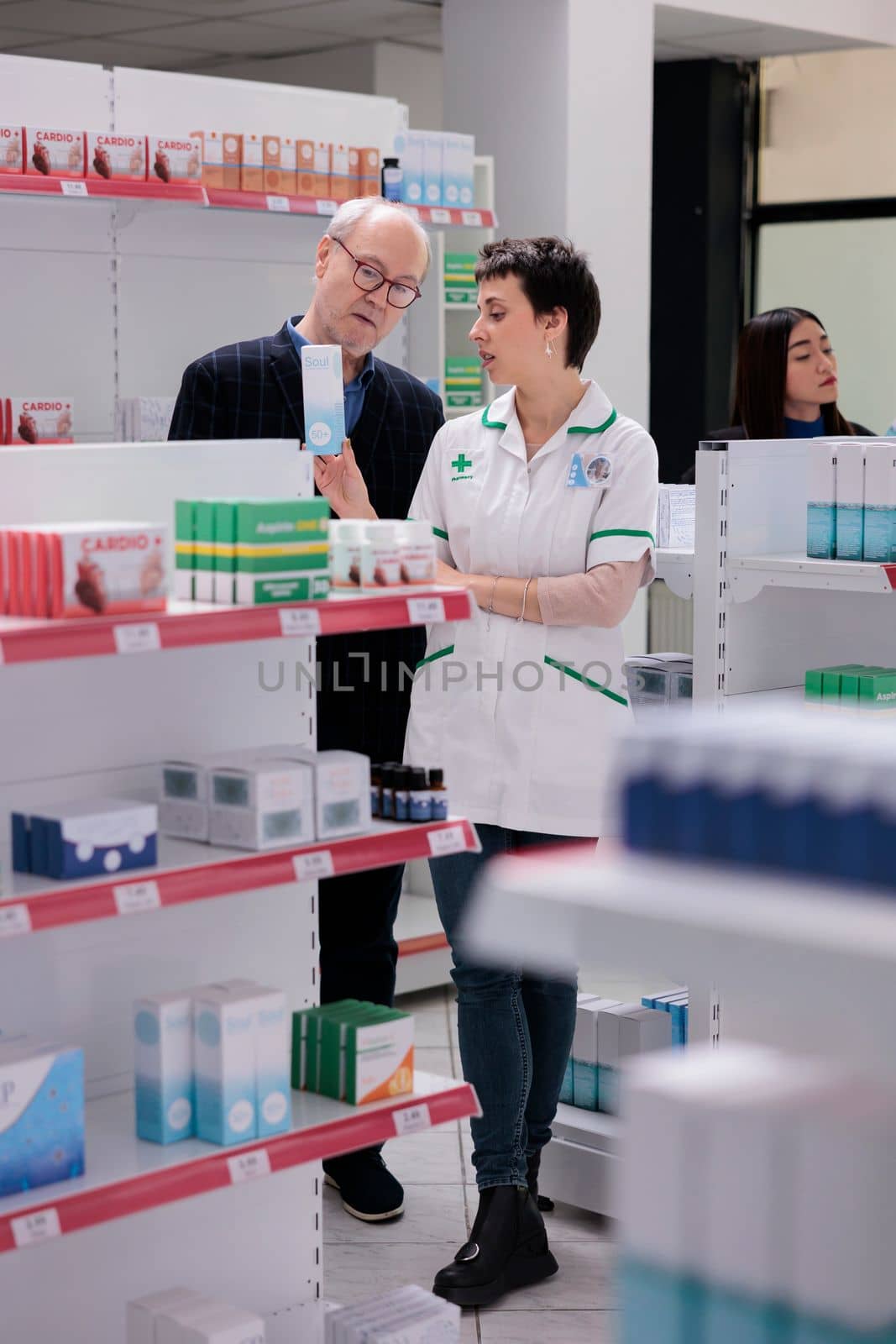 Pharmaceutical specialist holding sunscreen box and reading instruction to elderly buyer in drugstore. Old man listening to young pharmacy employee explaining sunblock lotion usage