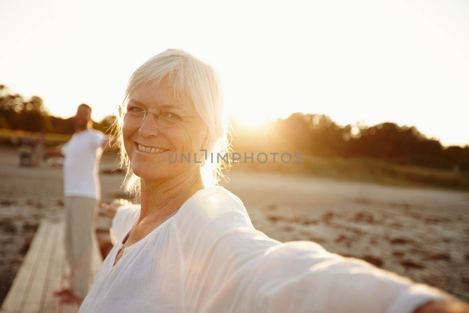 Yoga makes me feel great. a mature woman doing yoga on the beach with her husband in the background. by YuriArcurs