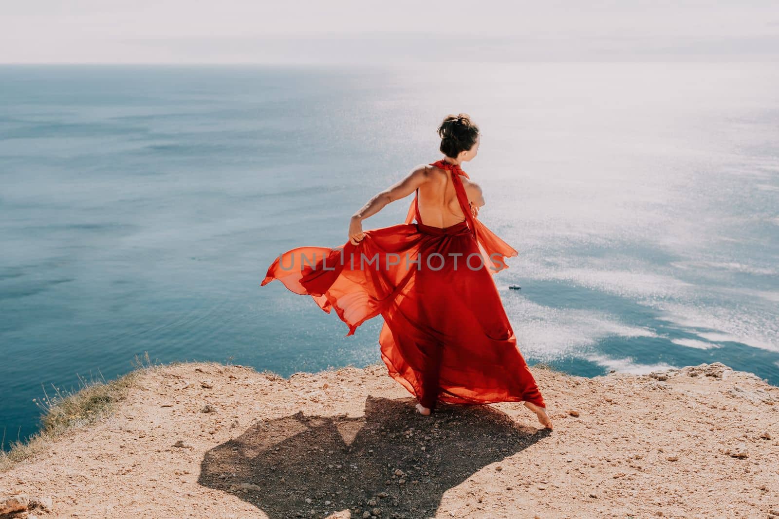 Side view a Young beautiful sensual woman in a red long dress posing on a rock high above the sea during sunrise. Girl on the nature on blue sky background. Fashion photo.