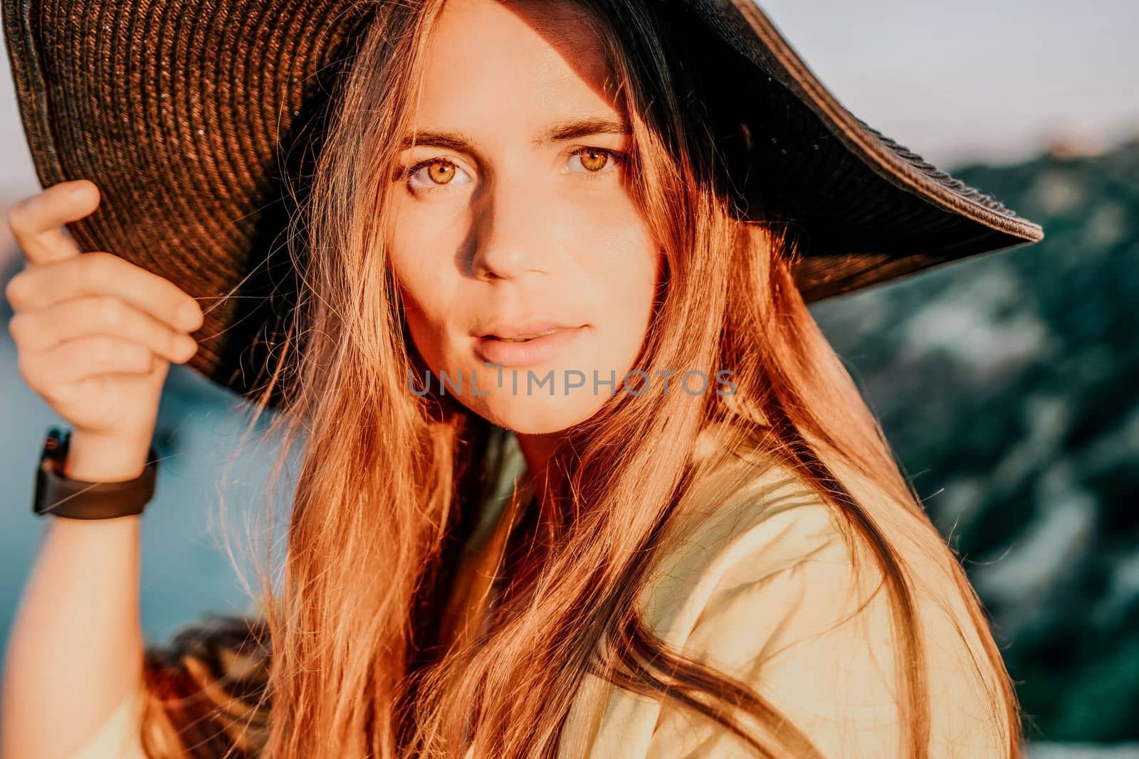 Portrait of happy young woman wearing summer black hat with large brim at beach on sunset. Closeup face of attractive girl with black straw hat. Happy young woman smiling and looking at camera at sea