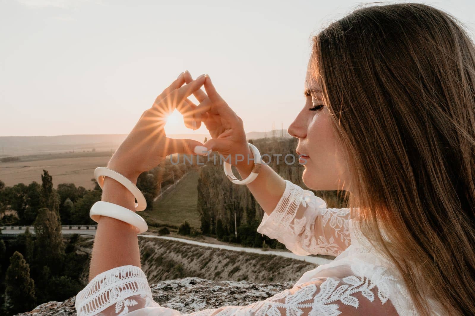 Happy woman in white boho dress making heart sign with hands on sunset in mountains. Romantic woman with long hair standing with her back on the sunset in nature in summer with open hands. by panophotograph