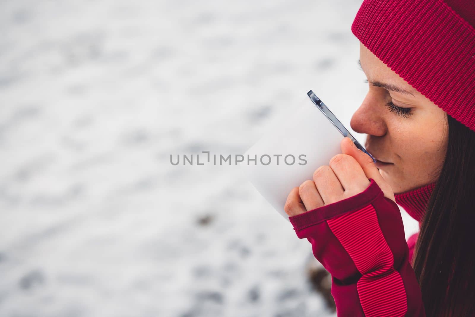 Beautiful young caucasian woman with brown hair, wearing a red jacket and a hat outside in the forest when it's snowing. Woman walking around a snowy forest. Woman holding a to go cup with tea. 