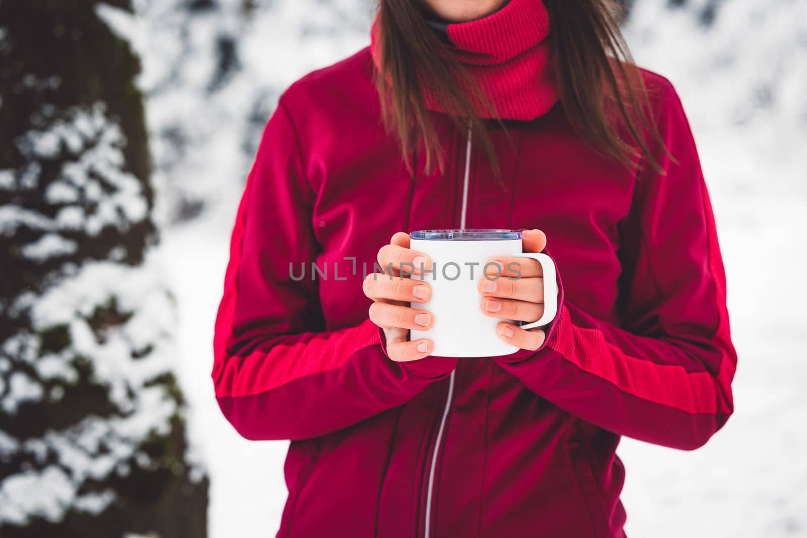 Beautiful young caucasian woman with brown hair, wearing a red jacket and a hat outside in the forest when it's snowing. Woman walking around a snowy forest. Woman holding a to go cup with tea. 