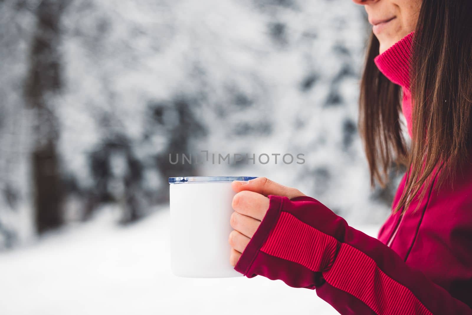 Beautiful young caucasian woman with brown hair, wearing a red jacket and a hat outside in the forest when it's snowing. Woman walking around a snowy forest. Woman holding a to go cup with tea. 