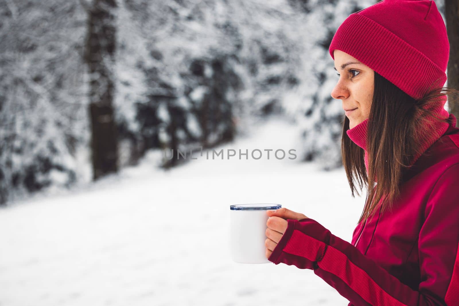 Beautiful young caucasian woman with brown hair, wearing a red jacket and a hat outside in the forest when it's snowing. Woman walking around a snowy forest. Woman holding a to go cup with tea. 