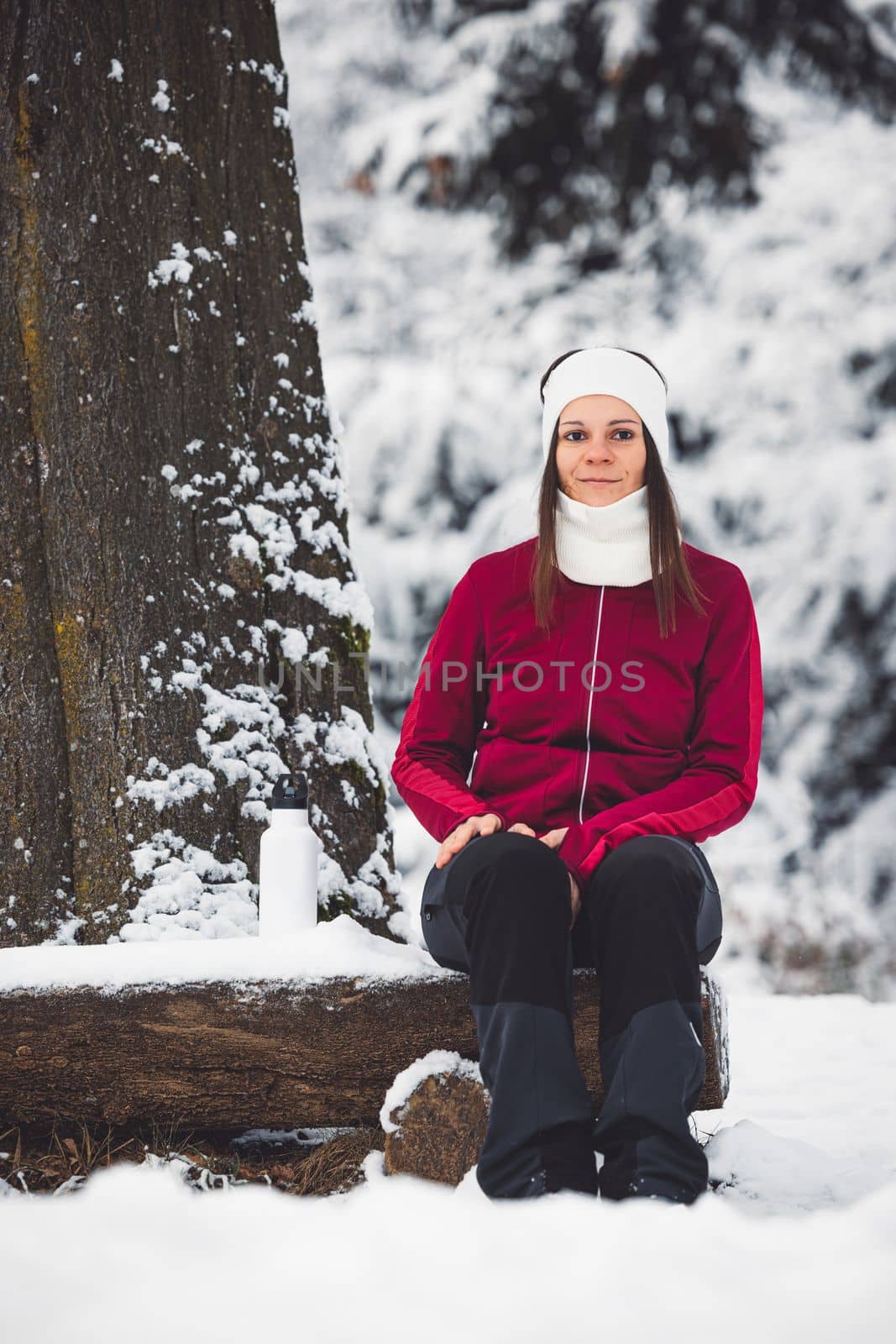 Beautiful young caucasian woman with brown hair, wearing a red jacket and a hat outside in the forest when it's snowing. Woman walking around a snowy forest. Woman holding a to go cup with tea. 