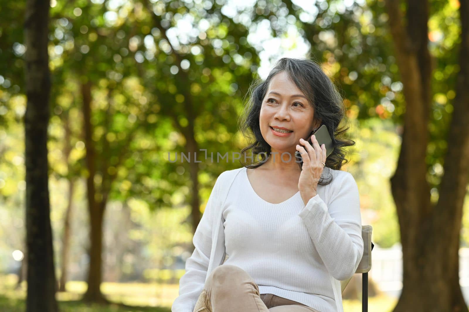 Pleased middle age woman talking on mobile phone while sitting in public park surrounded by green trees at sunlight morning.