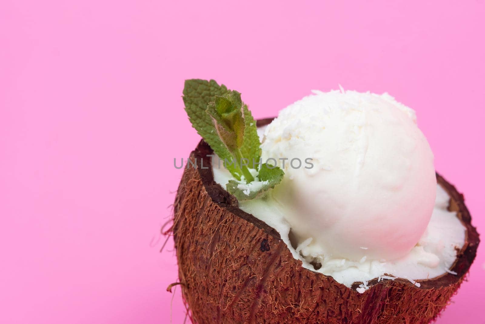 Vanilla ice cream balls in fresh coconut halves, decorated with mint leaves on a pink background.