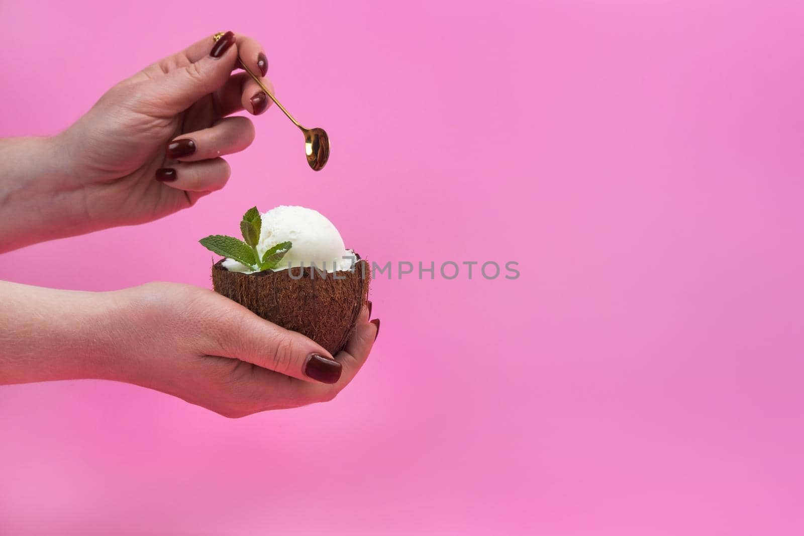 Vanilla ice cream balls in a fresh coconut half, decorated with mint leaves in the hand with a spoon on a pink background.