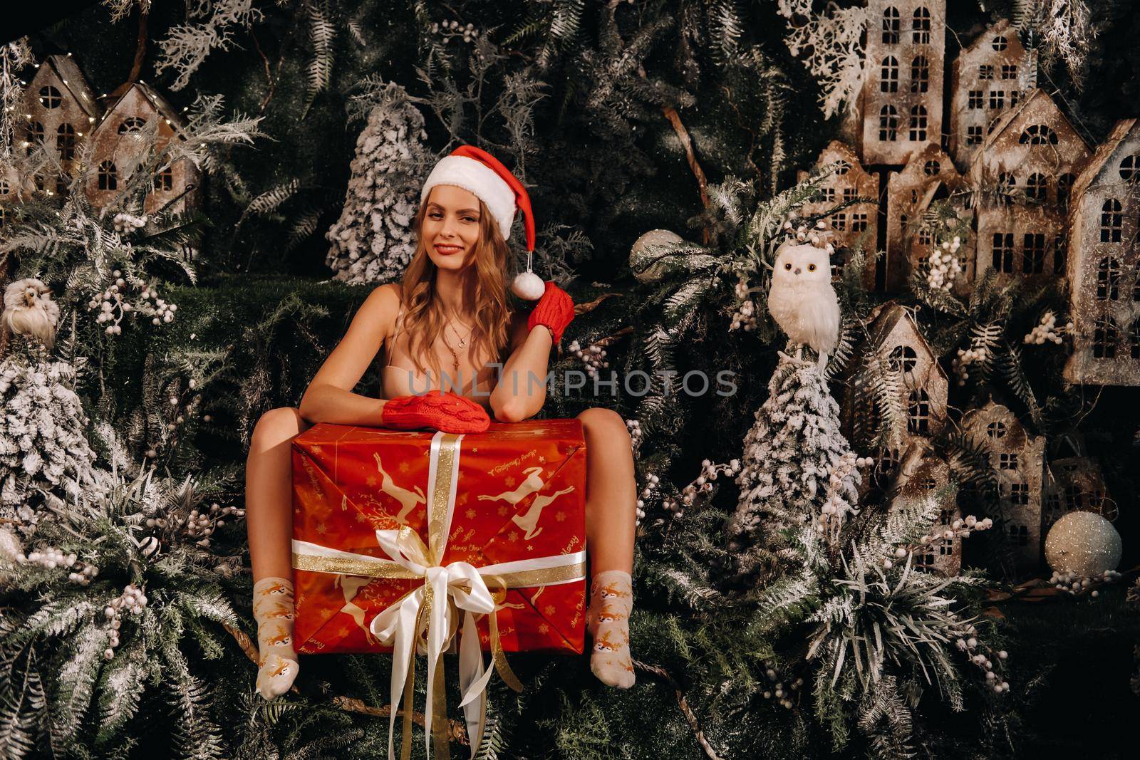 a girl in a Santa hat with a big Christmas gift in her hands on a fabulous Christmas background.Smiling woman in a bathing suit on the background of Christmas trees and small houses by Lobachad