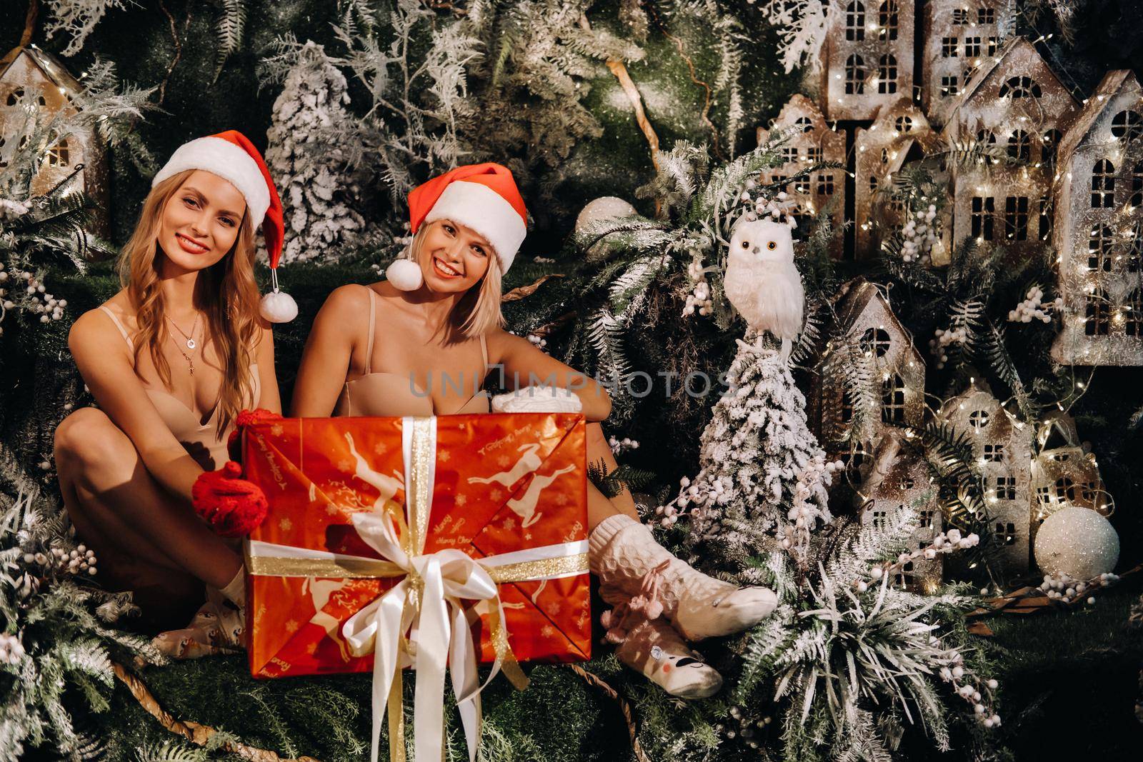 girls in Santa Claus hats with a big Christmas gift in their hands on a fabulous Christmas background.Smiling women in swimsuits on the background of Christmas trees and small houses.