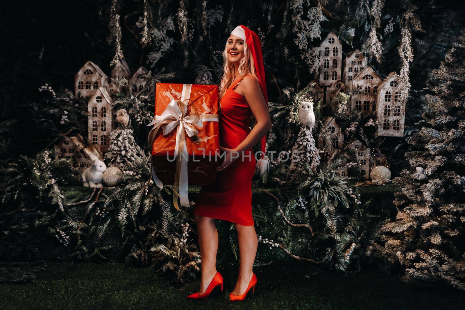 a girl in a Santa hat with a big Christmas gift in her hands on a fabulous Christmas background.A smiling woman in a red dress on the background of Christmas trees and small houses by Lobachad