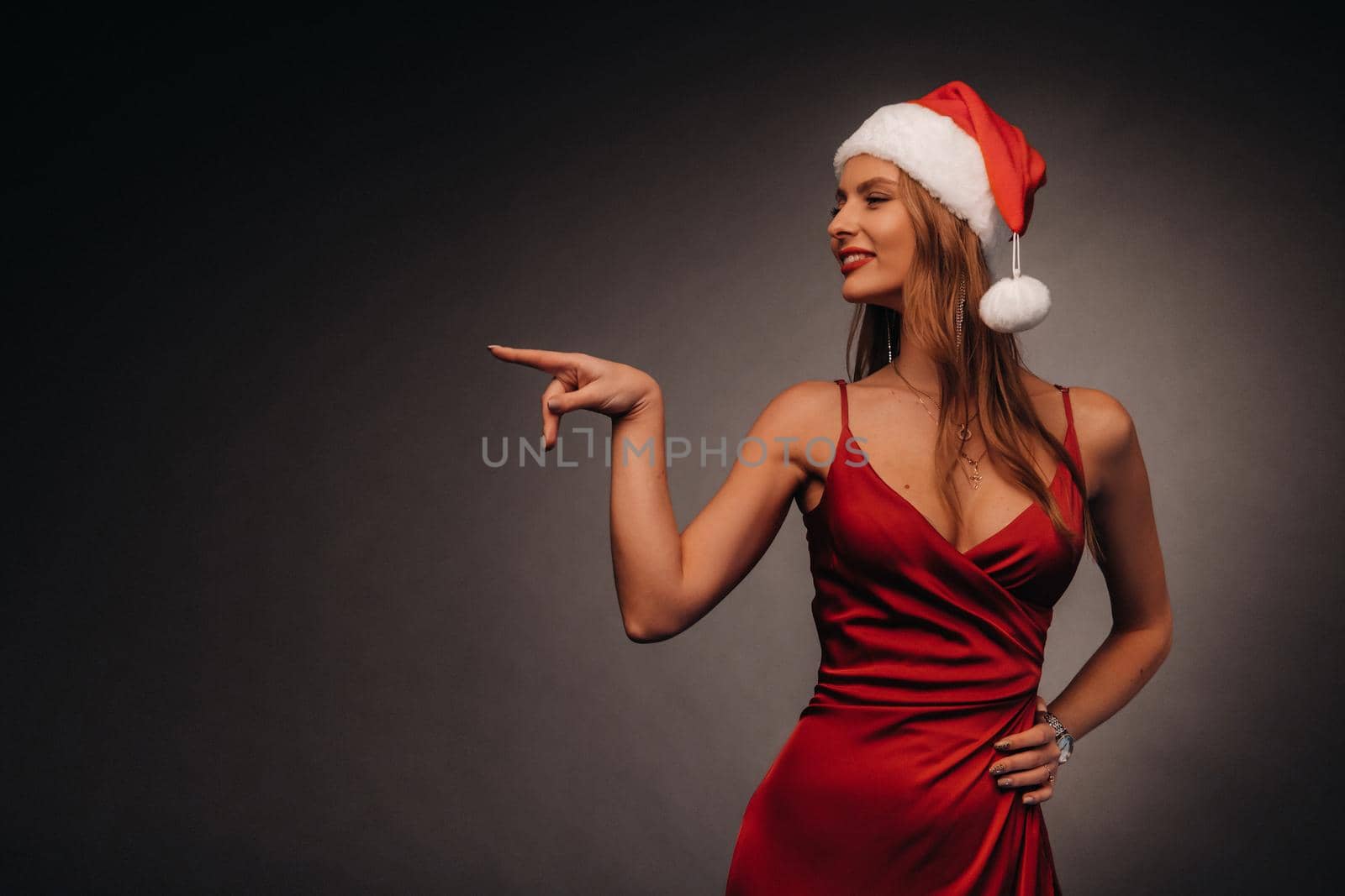 a woman in a red dress and a Christmas hat points her fingers at a black background.Smiling girl in new year shows direction on dark background by Lobachad