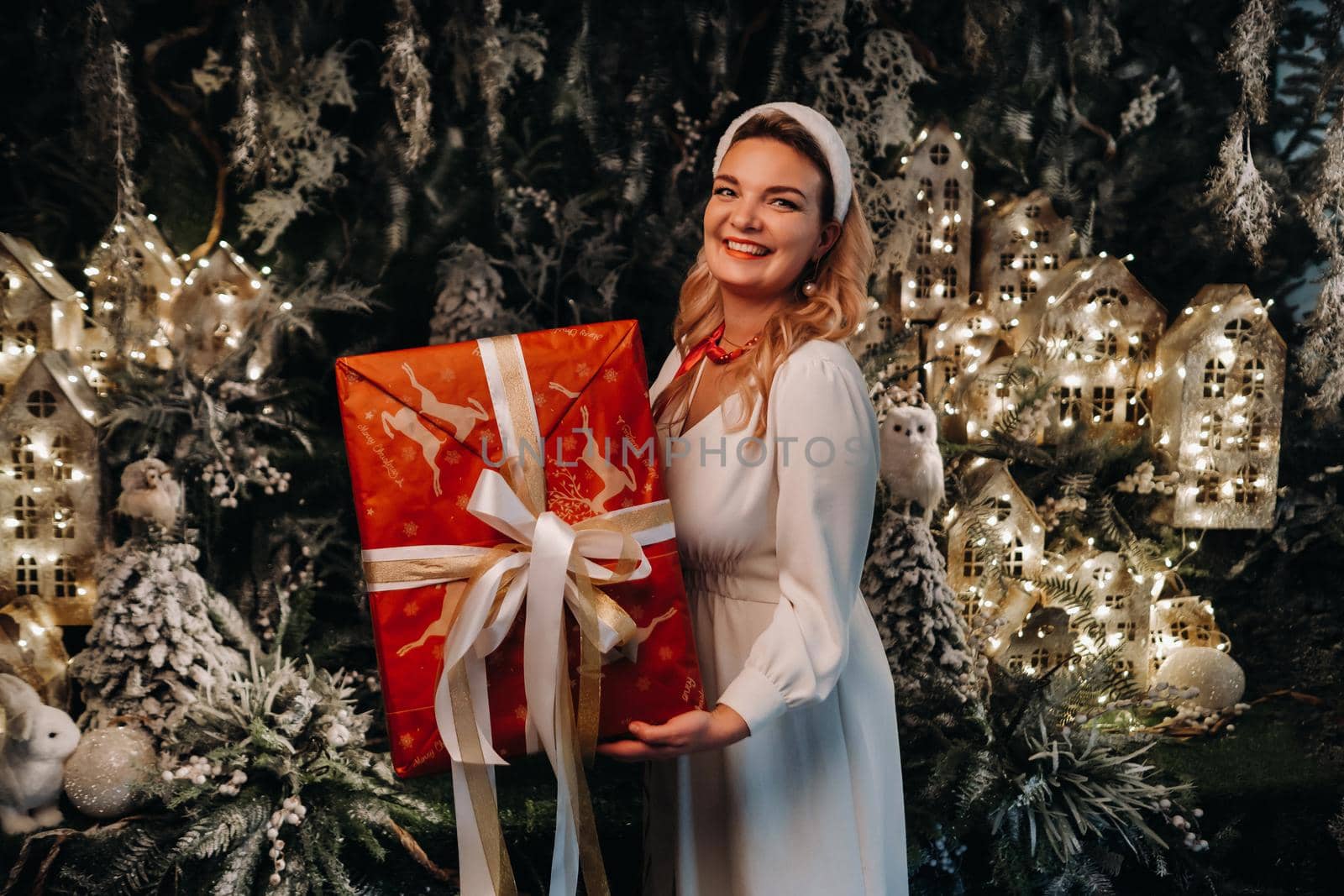 a girl in a white dress with a large Christmas gift in her hands on a fairy-tale background.Smiling woman in white clothes on the background of Christmas trees and small houses.