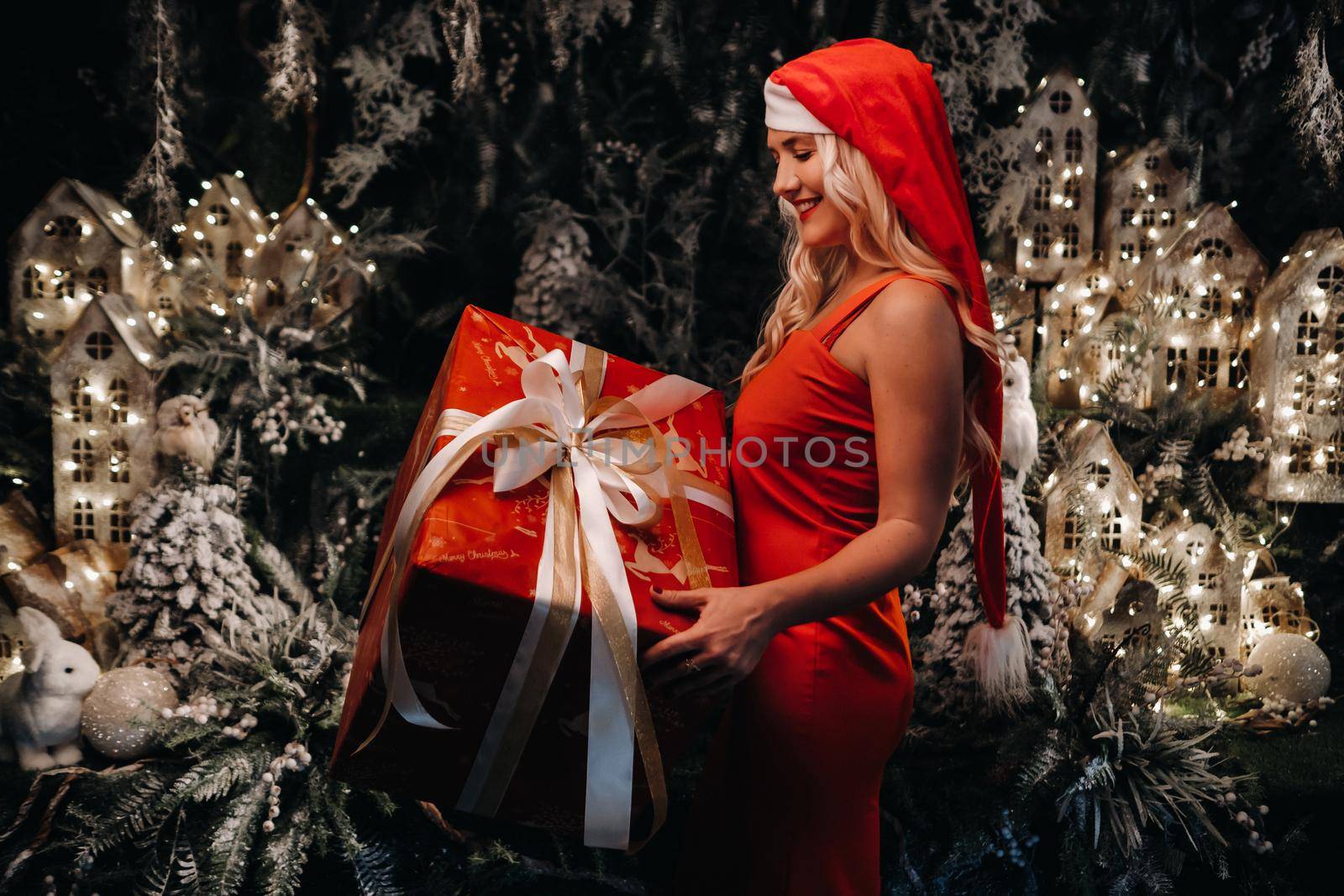 a girl in a Santa hat with a big Christmas gift in her hands on a fabulous Christmas background.A smiling woman in a red dress on the background of Christmas trees and small houses.