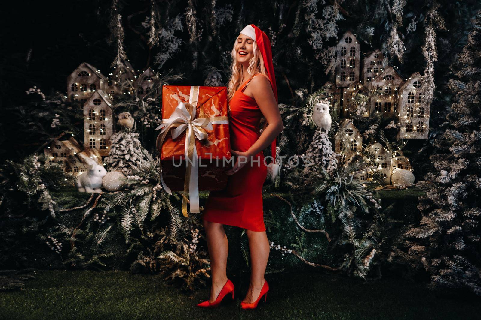 a girl in a Santa hat with a big Christmas gift in her hands on a fabulous Christmas background.A smiling woman in a red dress on the background of Christmas trees and small houses.