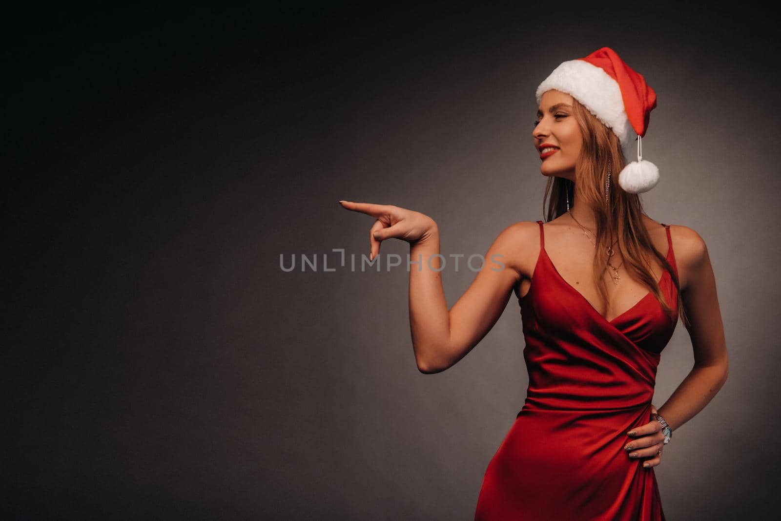 a woman in a red dress and a Christmas hat points her fingers at a black background.Smiling girl in new year shows direction on dark background.