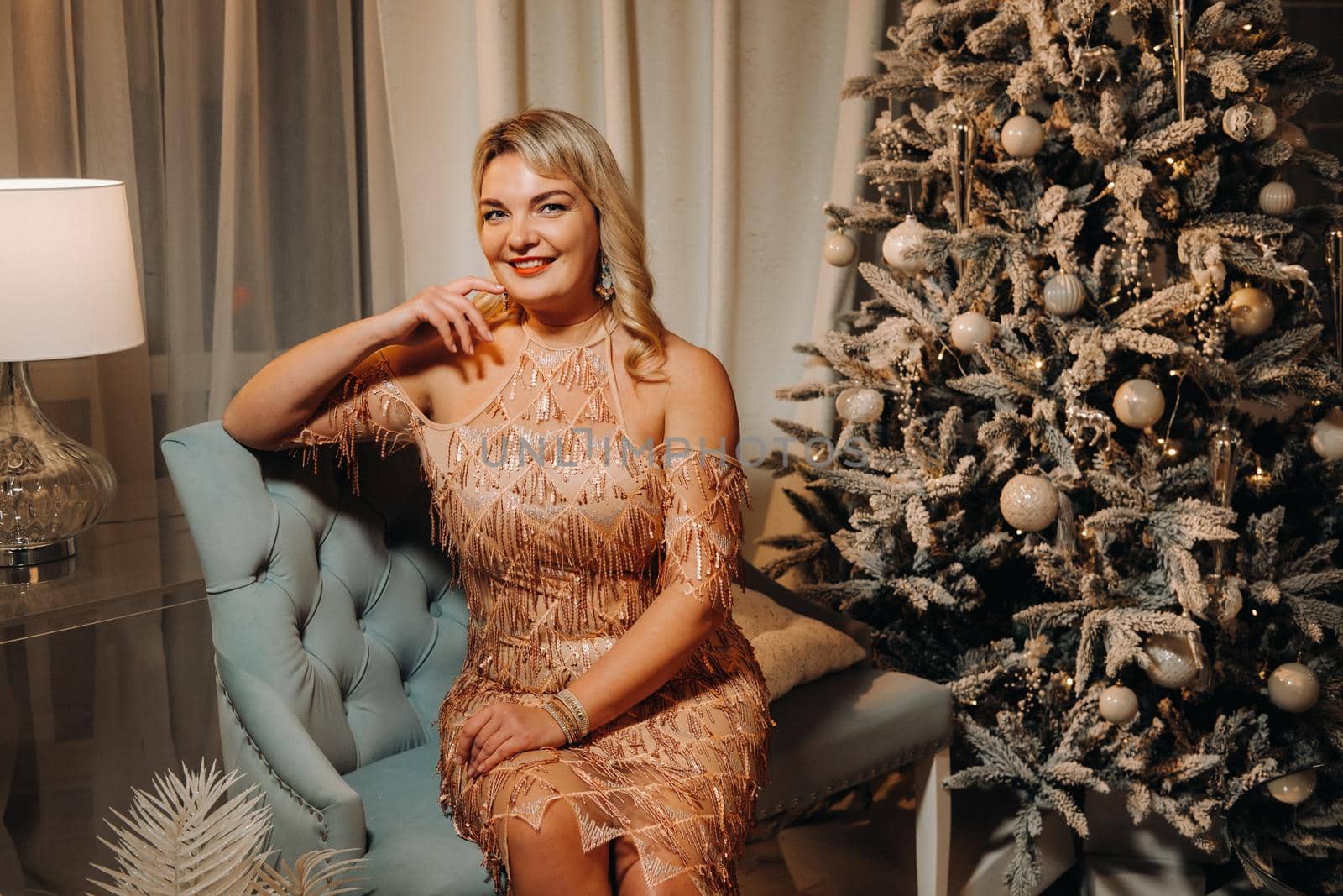 A girl in an evening dress is sitting on the sofa near the Christmas tree.