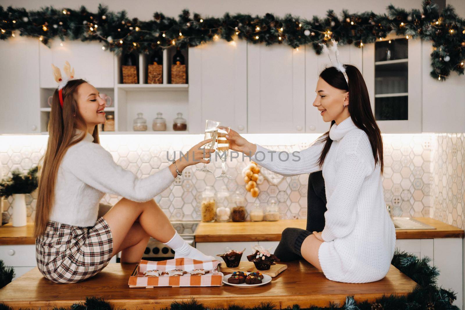 two girls in a cozy home environment in the kitchen with champagne in their hands for Christmas. Smiling girls drink champagne on a festive evening by Lobachad