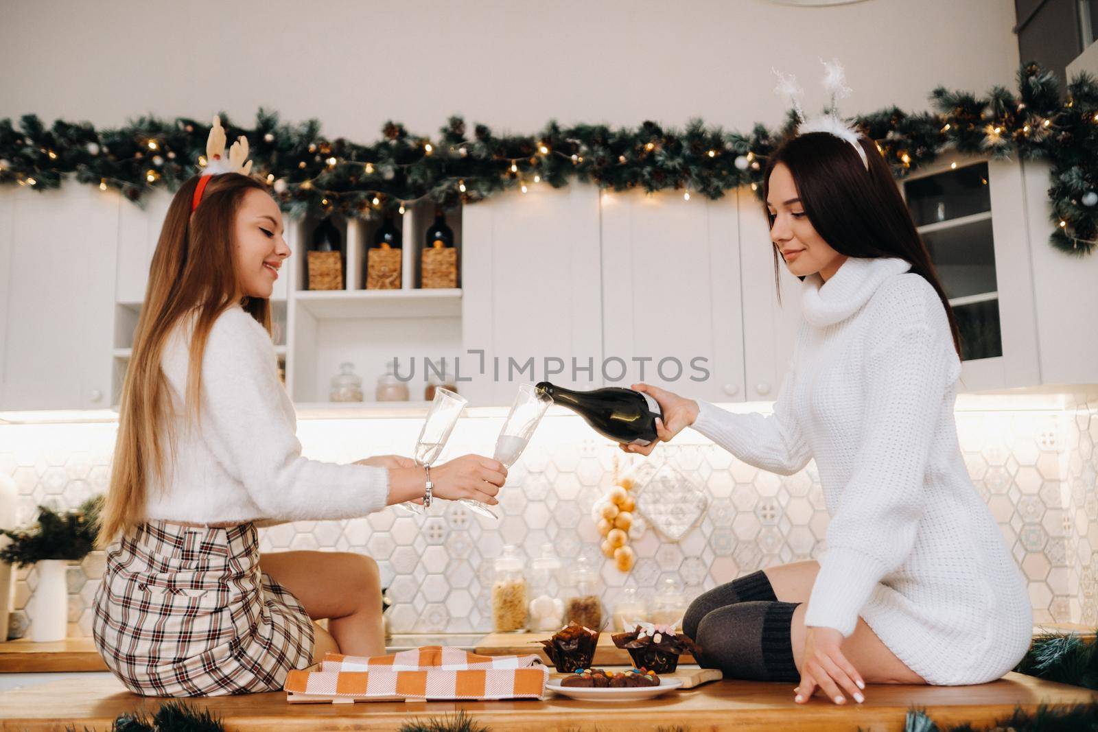 two girls in a cozy home environment in the kitchen pour champagne for Christmas. Smiling girls drink champagne on a festive evening.
