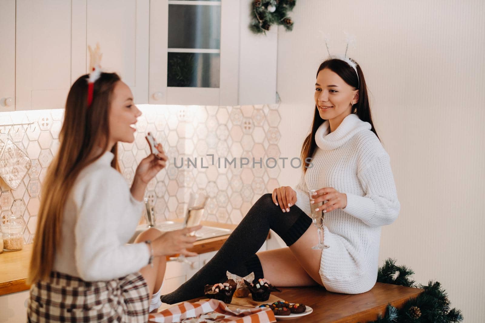 two girls in a cozy home environment in the kitchen with champagne in their hands for Christmas. Smiling girls drink champagne on a festive evening.