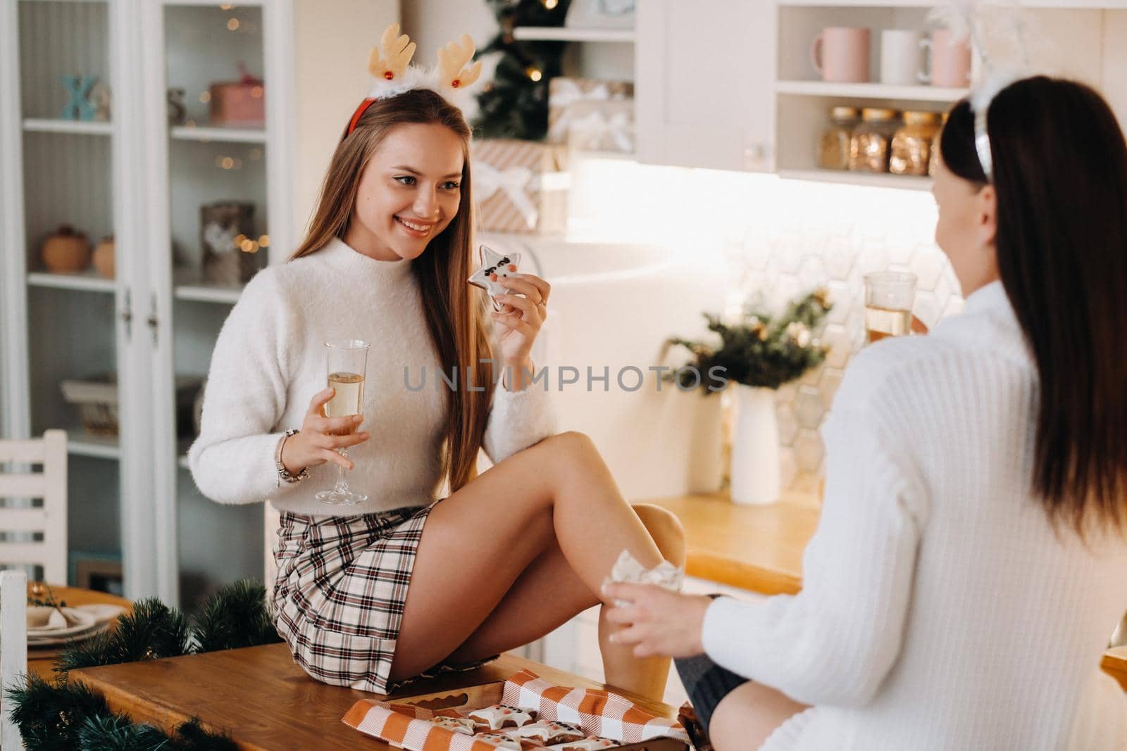 two girls in a cozy home environment in the kitchen with champagne in their hands for Christmas. Smiling girls drink champagne on a festive evening.