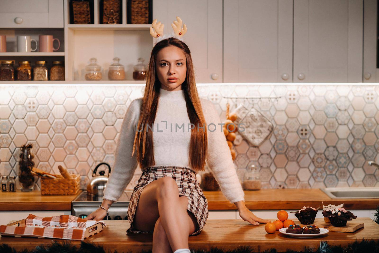 A girl with reindeer horns at Christmas sits on the kitchen table and smiles.Woman in new year's in the kitchen by Lobachad