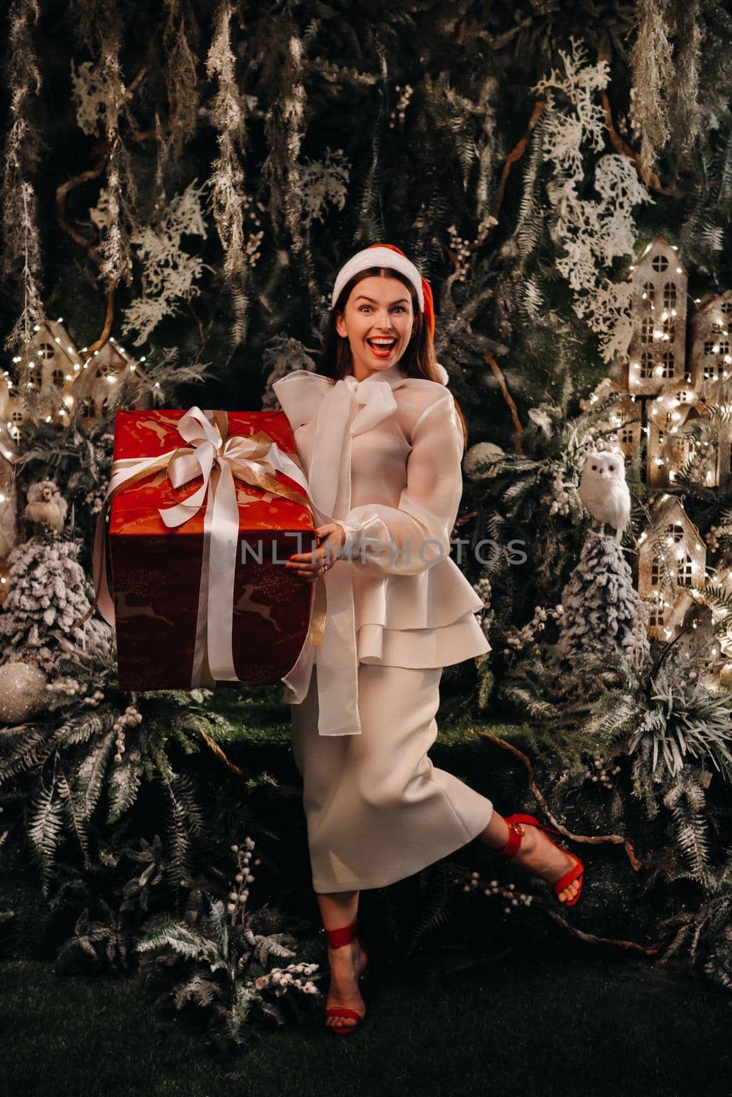 a girl in a Santa hat with a big Christmas gift in her hands on a fairy-tale island.Smiling woman in white clothes on the background of Christmas trees and small houses.