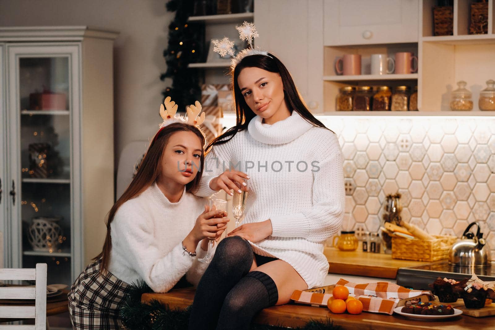 two girls in a cozy home environment in the kitchen with champagne in their hands for Christmas. Smiling girls drink champagne on a festive evening.