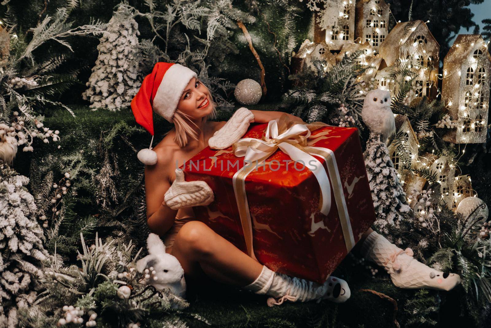 a girl in a Santa hat with a big Christmas gift in her hands on a fabulous Christmas background.Smiling woman in a bathing suit on the background of Christmas trees and small houses.