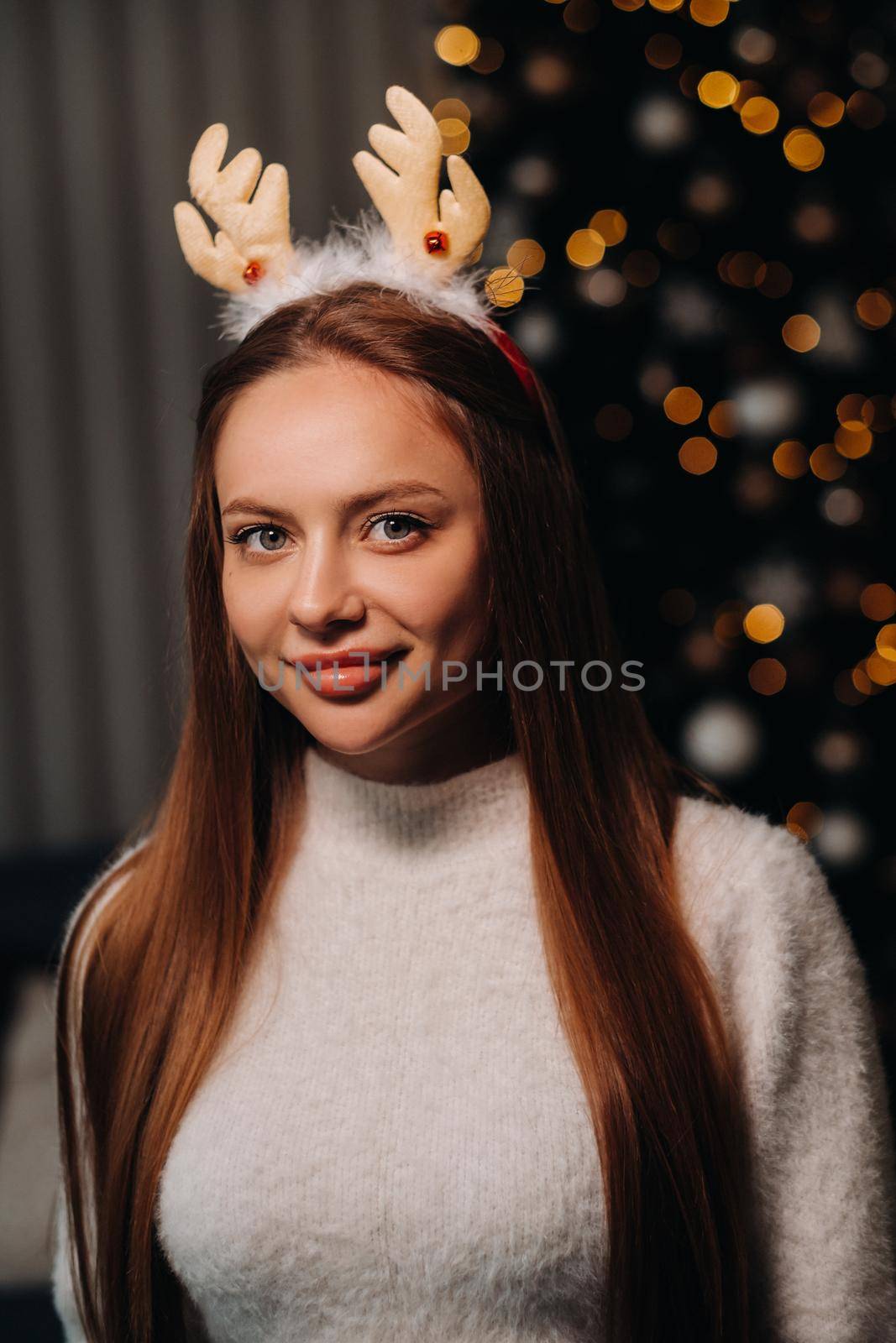 Girl for Christmas with reindeer horns in the home interior.A woman on New year's eve in a white sweater and skirt by Lobachad