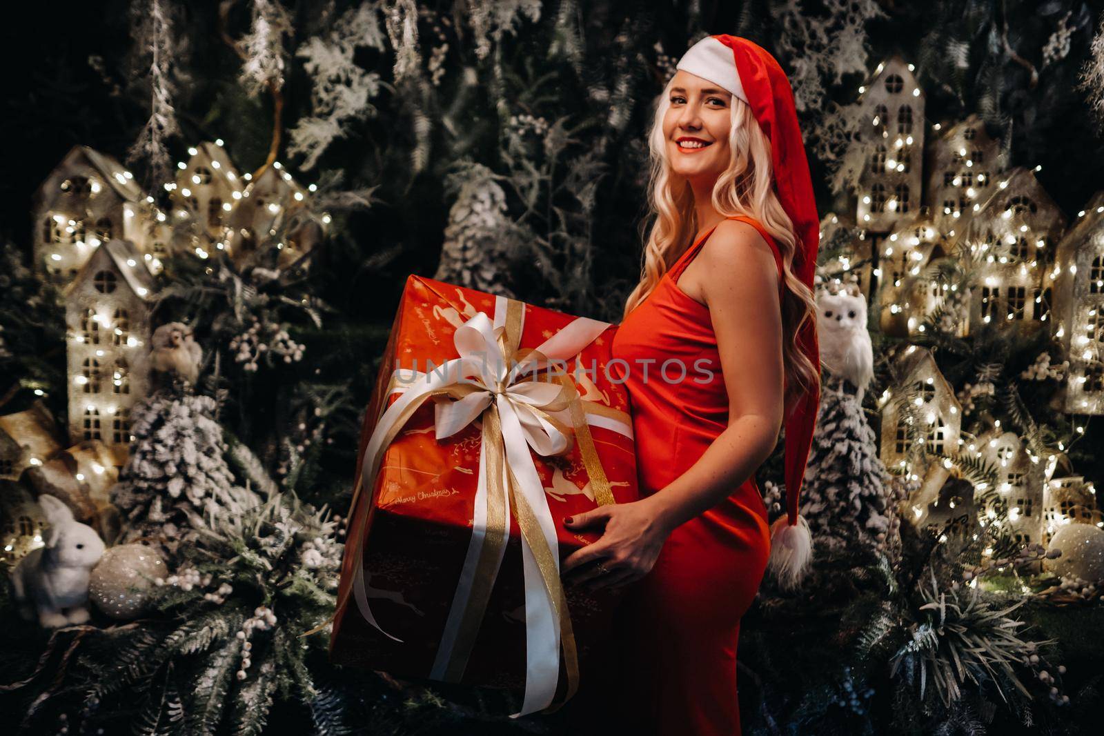 a girl in a Santa hat with a big Christmas gift in her hands on a fabulous Christmas background.A smiling woman in a red dress on the background of Christmas trees and small houses.