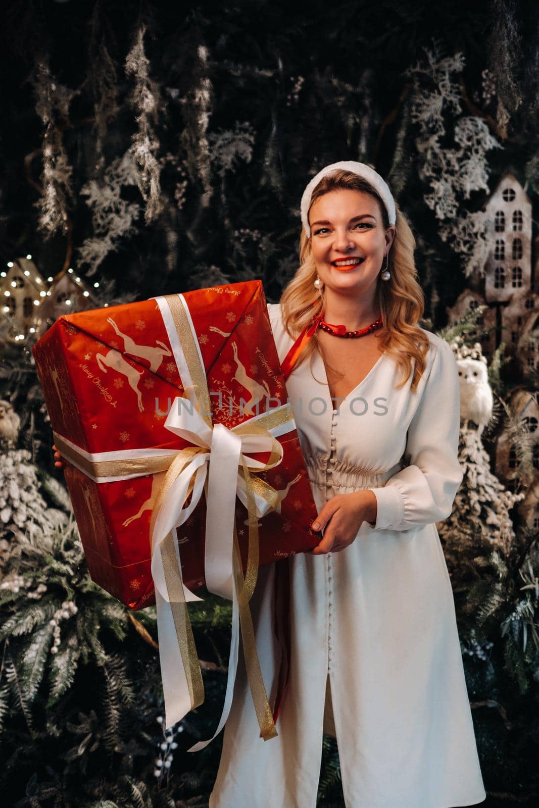 a girl in a white dress with a large Christmas gift in her hands on a fairy-tale background.Smiling woman in white clothes on the background of Christmas trees and small houses.