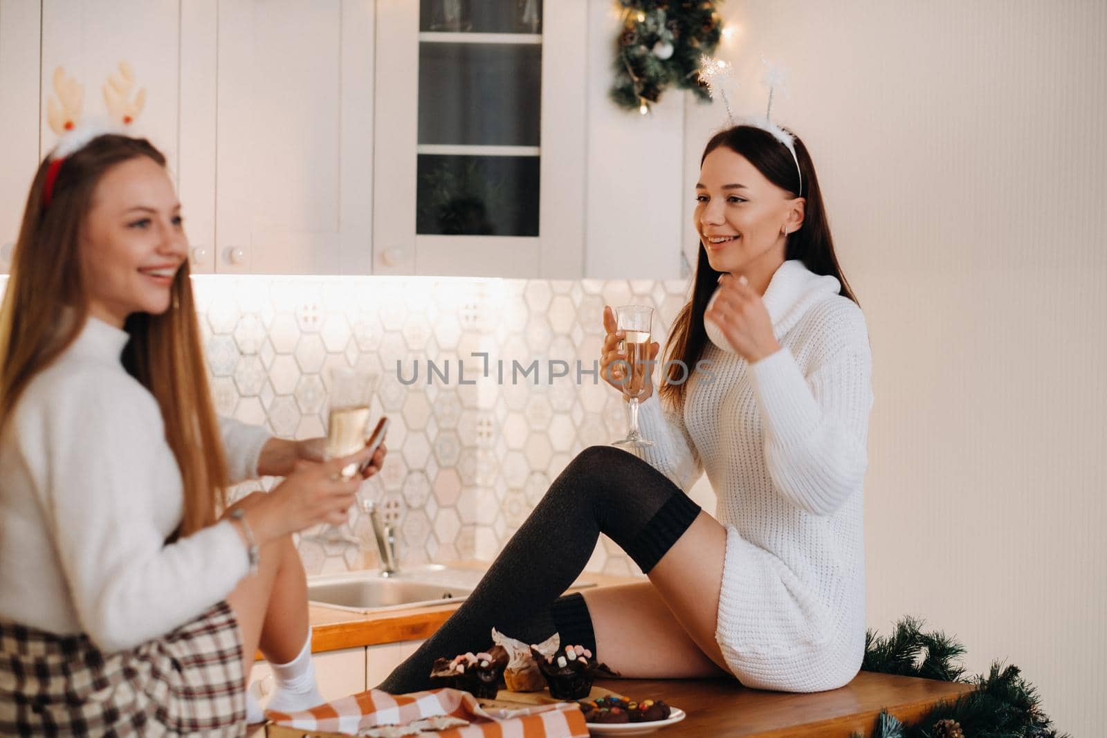 two girls in a cozy home environment in the kitchen with champagne in their hands for Christmas. Smiling girls drink champagne on a festive evening.