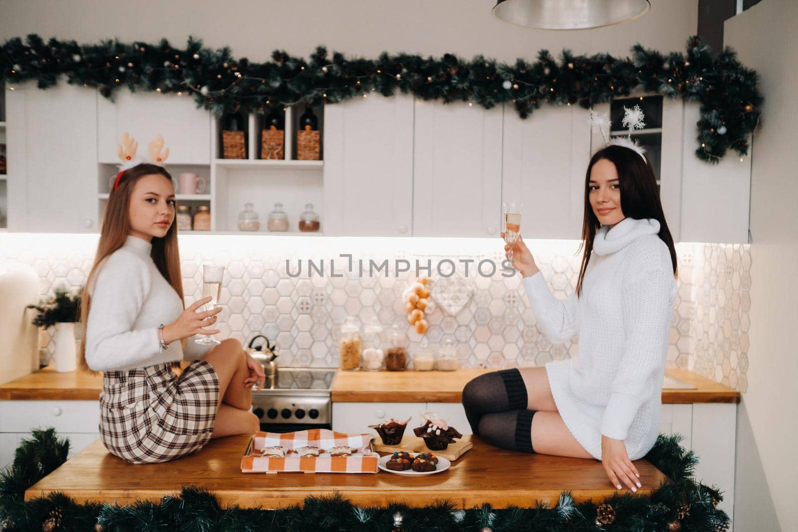 two girls in a cozy home environment in the kitchen with champagne in their hands for Christmas. Smiling girls drink champagne on a festive evening.