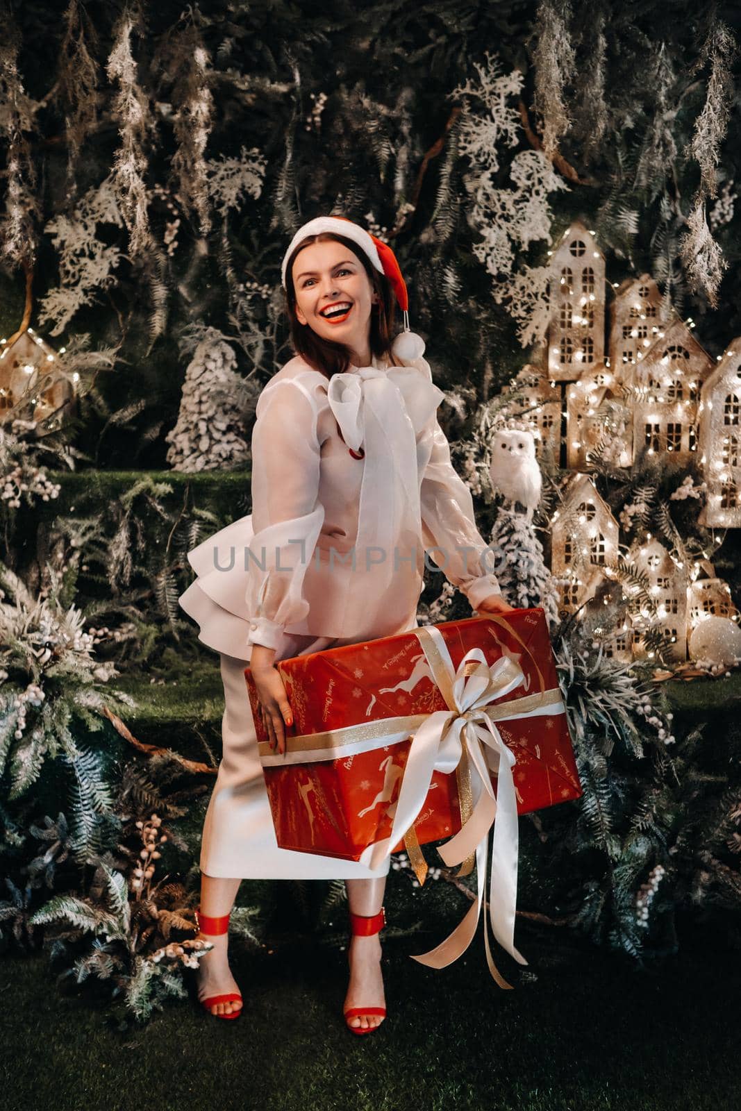 a girl in a Santa hat with a big Christmas gift in her hands on a fairy-tale island.Smiling woman in white clothes on the background of Christmas trees and small houses.