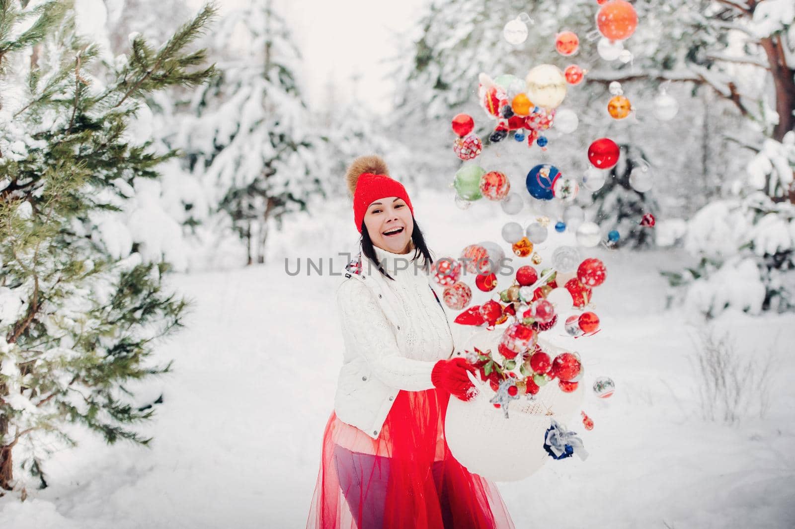 A girl in a white jacket throws Christmas balls to decorate the Christmas tree.A girl throws Christmas decorations from a basket into the winter forest by Lobachad