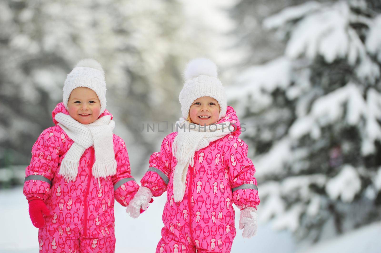 Two little twin girls in red suits stand in a snowy winter forest.