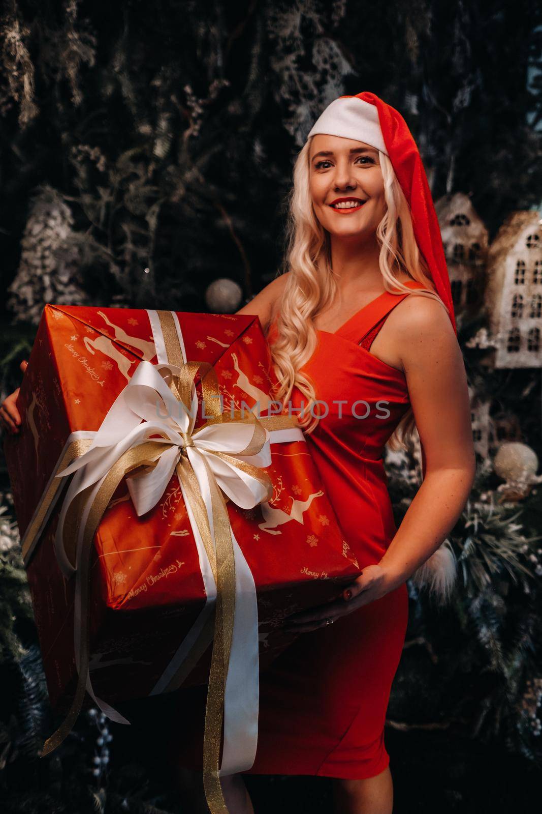 a girl in a Santa hat with a big Christmas gift in her hands on a fabulous Christmas background.A smiling woman in a red dress on the background of Christmas trees and small houses.