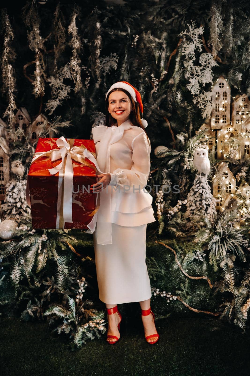 a girl in a Santa hat with a big Christmas gift in her hands on a fairy-tale island.Smiling woman in white clothes on the background of Christmas trees and small houses by Lobachad