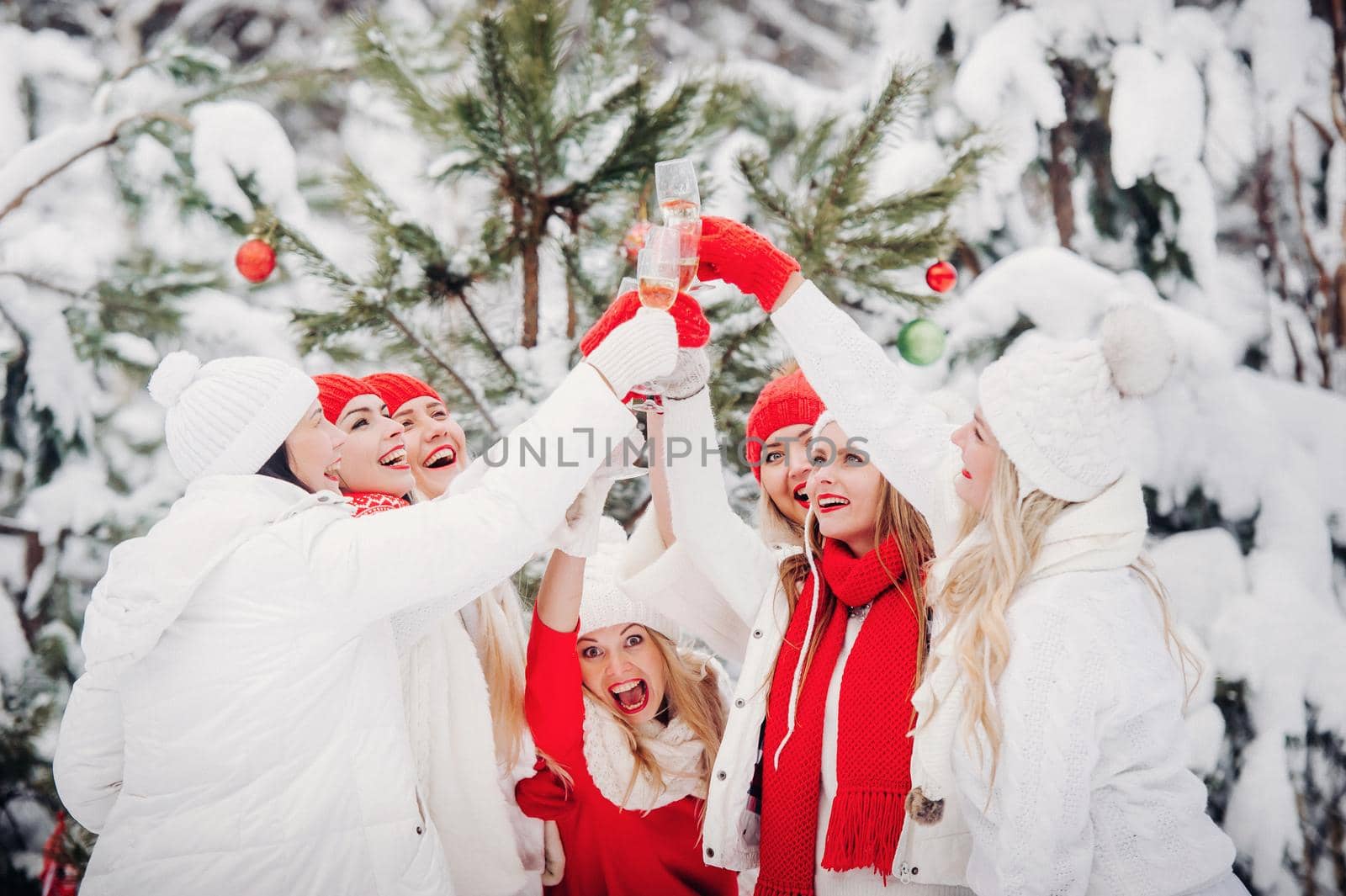 A large group of girls with glasses of champagne in their hands stands in the winter forest.Girls in red and white clothes with new year's drinks in a snow-covered forest