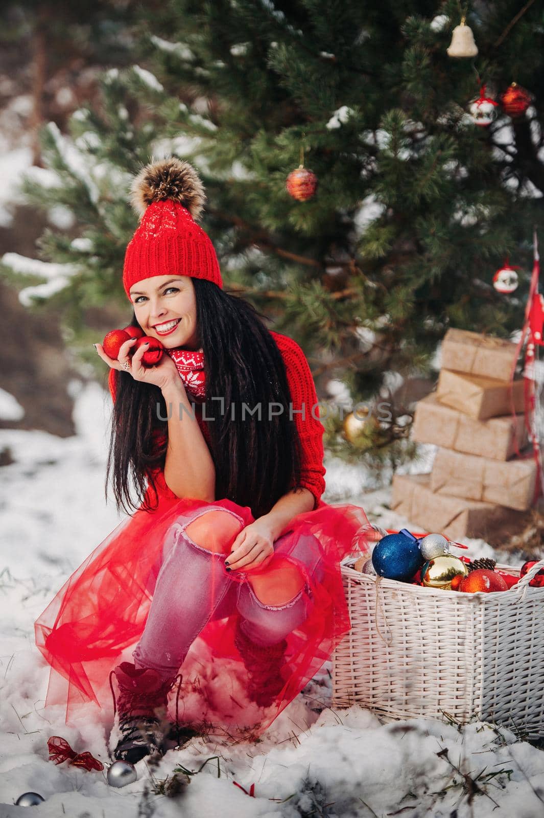 A girl in red clothes with Christmas balloons in her hands is sitting in a snow-covered forest.Woman with Christmas balls on the street in winter near the Christmas tree.